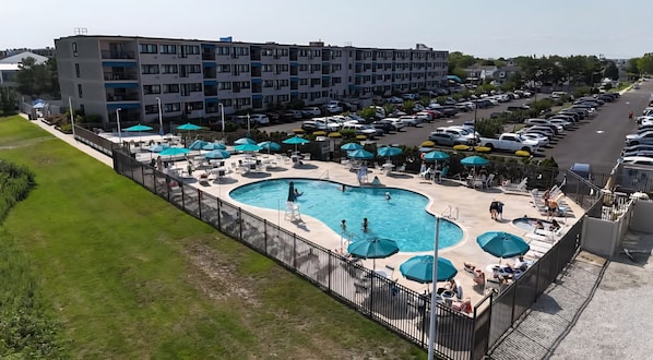 A swimming pool with multiple lanes, surrounded by lounge chairs and umbrellas, adjacent to a parking lot and a multi-story building. - ShoreLine | 2BR Coastal Condo + Pool (Brigantine)