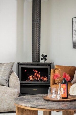 A living room with a wood-burning stove, a sofa, a coffee table with glasses and a bottle, and a wall-mounted fan. - Pepper Grove - Beautifully styled Margaret River retreat (Margaret River)