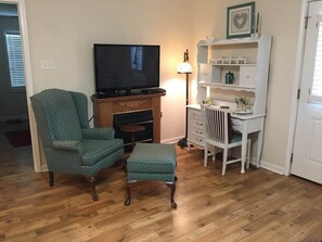 A living room with a television, a fireplace, a green armchair, a matching ottoman, a white dresser, and a lamp.