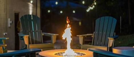 A round outdoor fire pit with a flame, surrounded by Adirondack chairs.