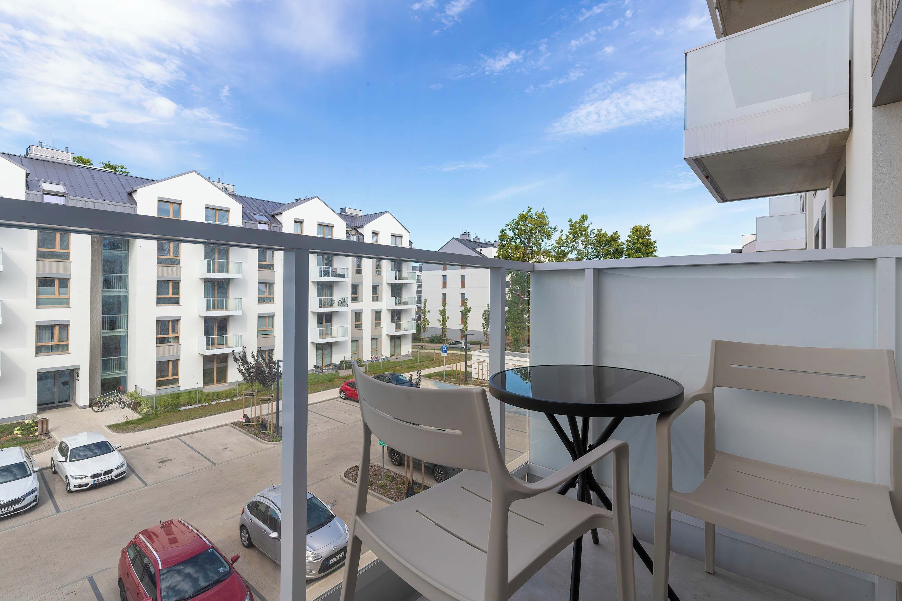 A balcony with a table and chairs, overlooking a parking area and residential buildings.