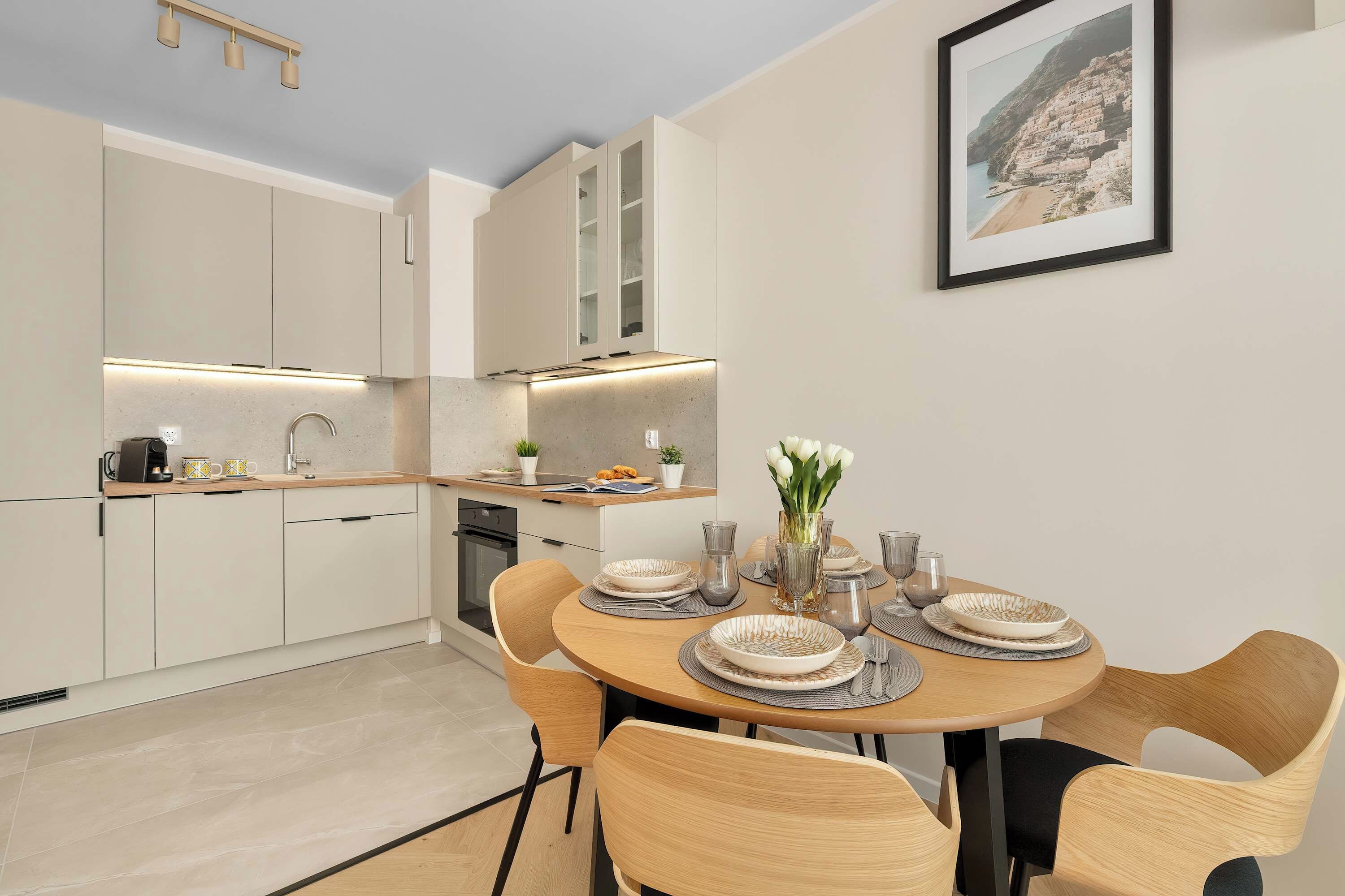 Una cocina moderna con electrodomésticos integrados, un comedor con una mesa redonda y sillas, y una fotografía enmarcada de la costa en la pared.