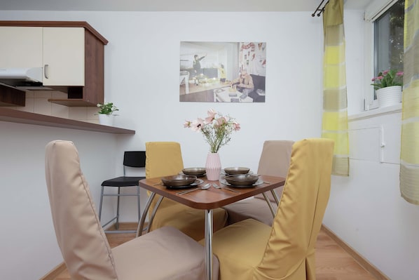 Ein Essbereich mit Tisch für zwei Personen, Stühlen, einer Vase mit Blumen und einer Küchenzeile mit Schränken und einem Fenster.