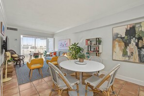 A dining area with a round table and wicker chairs, a bookshelf with books and plants, and a large abstract painting on the wall. - Serene 1-bedroom condo in beautiful Key Biscayne, 3 minute walk to the beach (Key Biscayne)