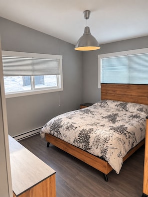 A bedroom with a bed, a nightstand, a wooden headboard, a pendant light, and a window with blinds. - Big Shell Lake Cabin | Sleeps 8 | Fireplace, Starlink & Private Yard (Shell Lake)