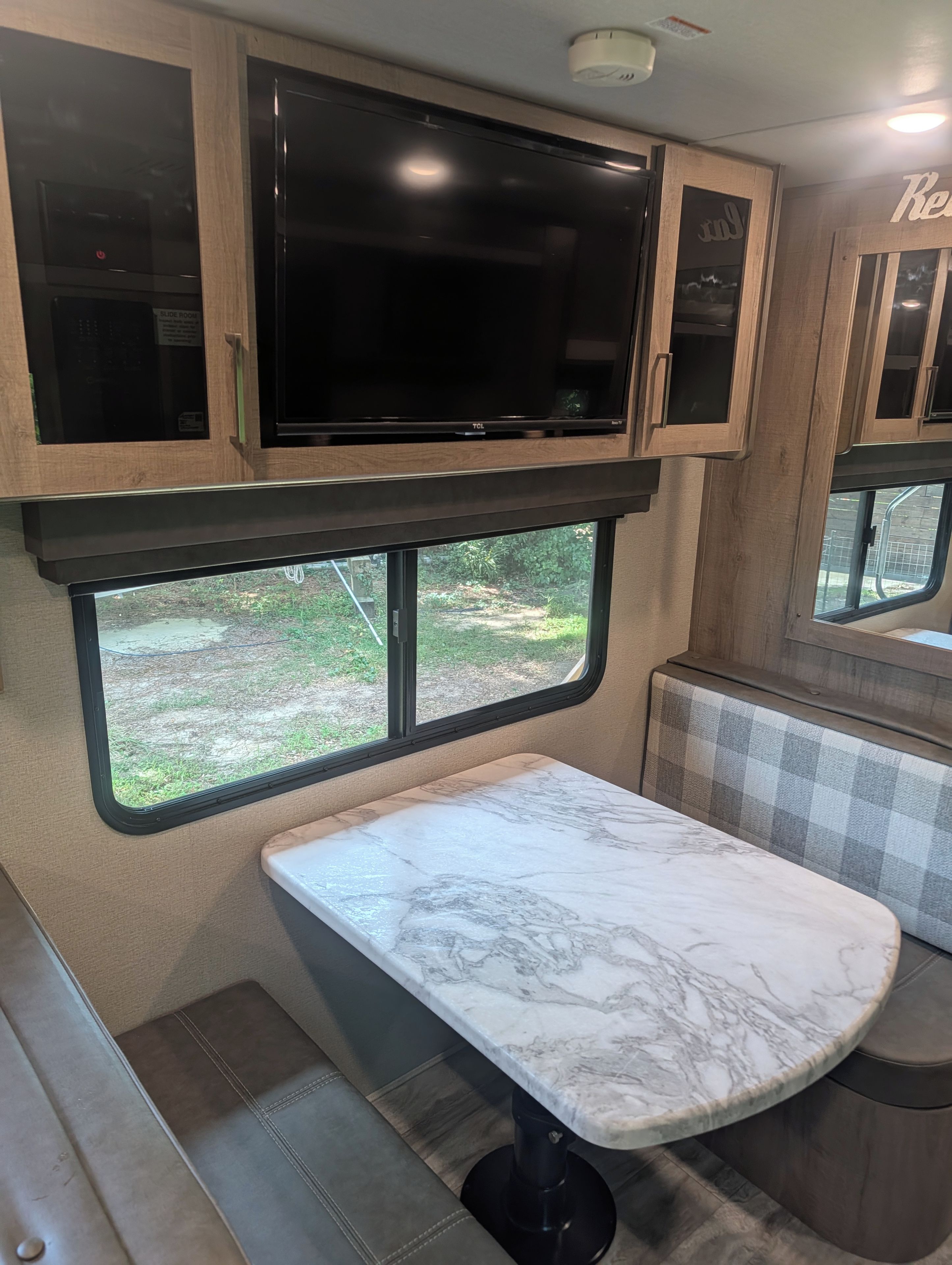A kitchen area with a marble countertop, a television above cabinets, and a window with a view of greenery.