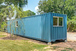 Een blauwe verzendcontainer met een raam, geplaatst op een grasveld met bomen op de achtergrond.