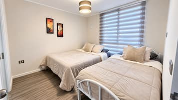 A bedroom with two beds, a window with blinds, and a wall-mounted light fixture.