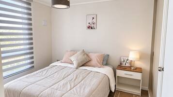 Quarto com cama, mesa de cabeceira, abajur e janela com persianas.