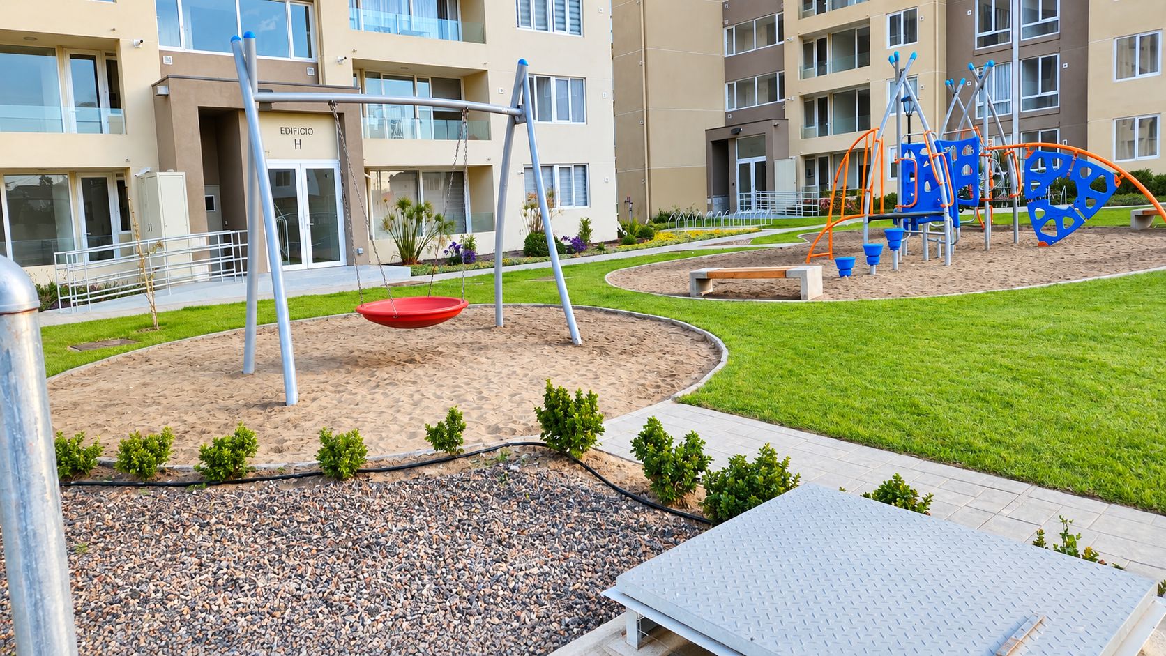 A playground with swings and climbing structures, surrounded by a landscaped area with small bushes and a paved path.