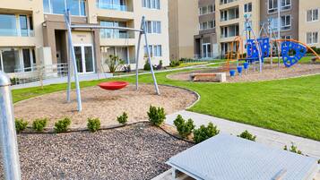 A playground with swings and climbing structures, surrounded by a landscaped area with small bushes and a paved path.