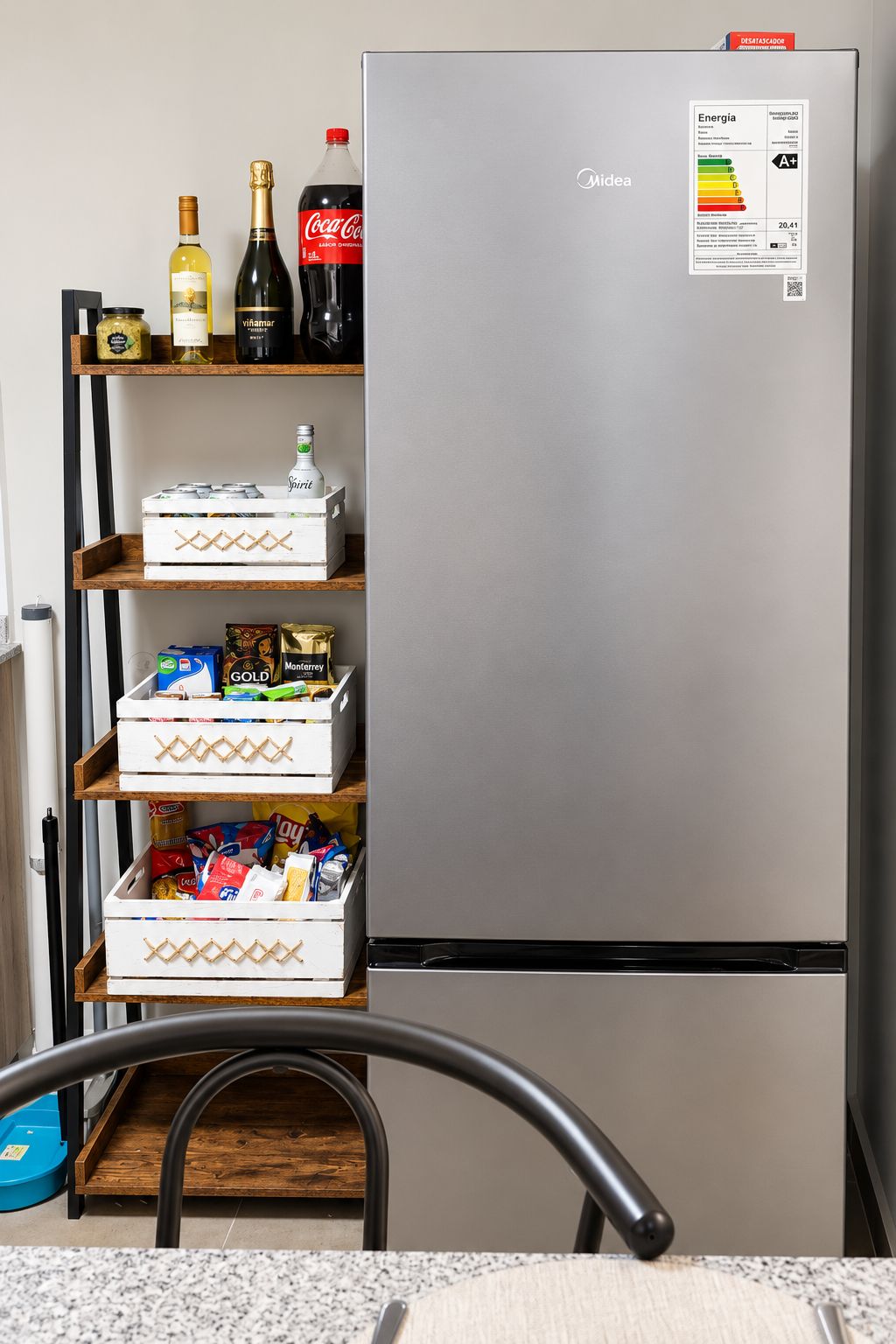 Geladeira de aço inoxidável com etiqueta energética Midea, prateleiras com alimentos e uma cadeira à frente.