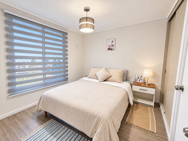 Un dormitorio con cama, mesita de noche, lámpara, ventana con persianas y una puerta.