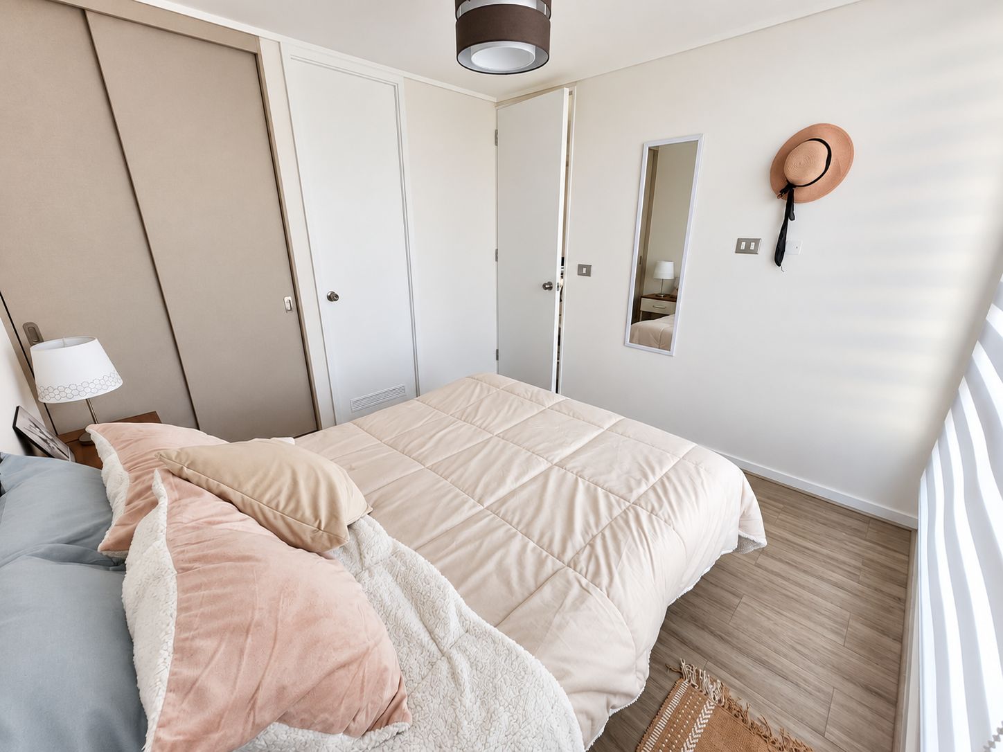 A bedroom with a bed, a nightstand, a lamp, a wardrobe, a mirror, and a hat on the wall.