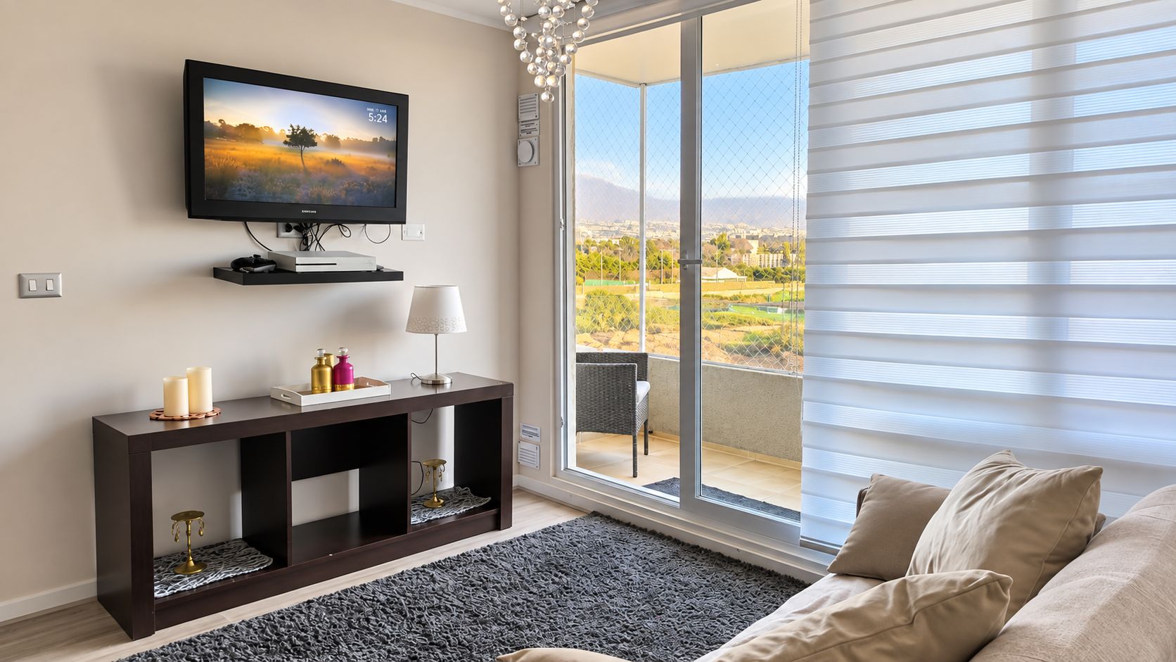 A living room with a TV, console table, and a sofa. There's a large window with blinds and a view of a landscape.