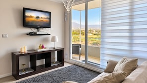 Una sala de estar con televisor, consola y sofá. Hay un ventanal con persianas y vistas al paisaje.