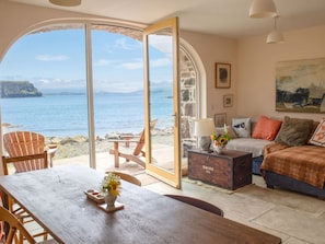 A room with a large window overlooking a body of water, a wooden table, a sofa, and a chest of drawers. - The Boat house by Interhome (North Skye)
