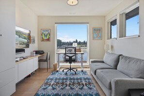 A modern living room with a grey sofa, a desk with a computer, and a large window offering a scenic view. - Gorgeous Deschutes Riverfront, 4-bedroom home in Old Mill/ Bend! (Bend)
