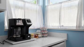 Una cocina con cafetera, un escurreplatos para tazas y una ventana con cortinas traslúcidas.