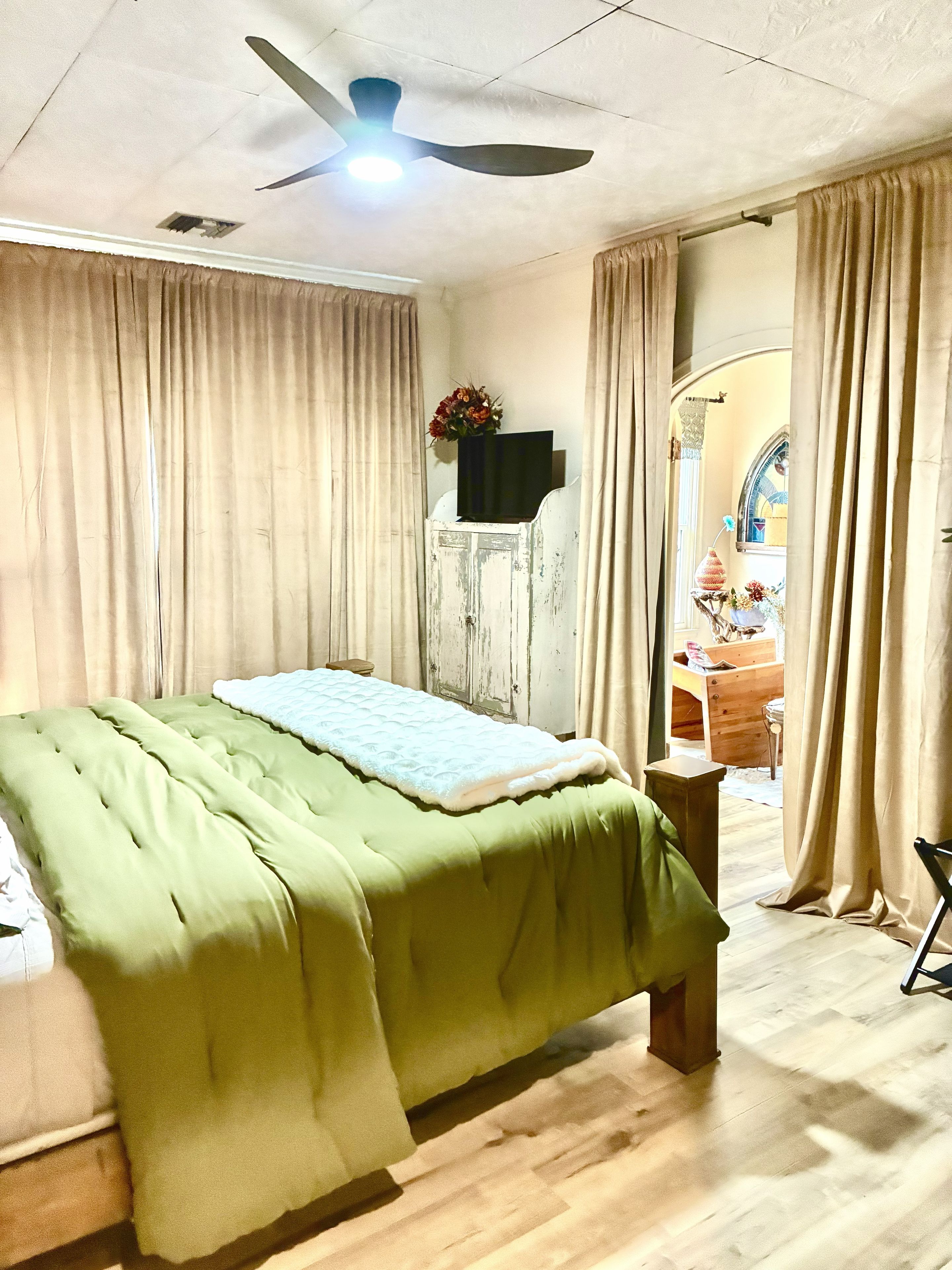 Een kamer met een bed, een plafondventilator, een televisie op een houten kast en uitzicht op een buitenruimte door een deuropening.