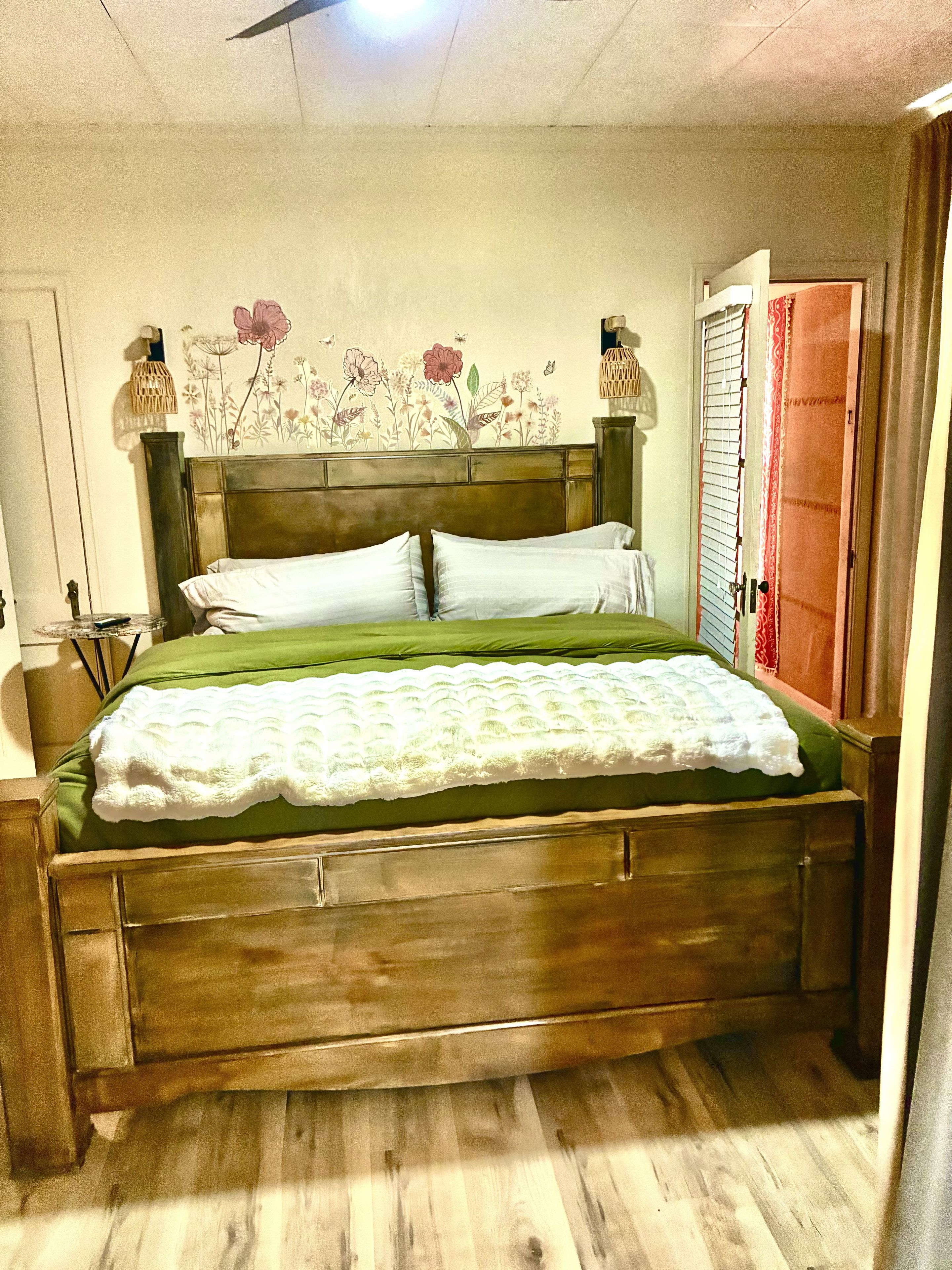 Una cama de madera con una colcha verde y almohadas blancas, un cabecero de madera, una pared con papel tapiz floral y un armario de madera con una cortina roja.