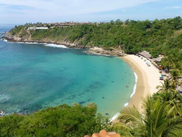 A coastal view with a beach, clear turquoise waters, and a forested hillside.