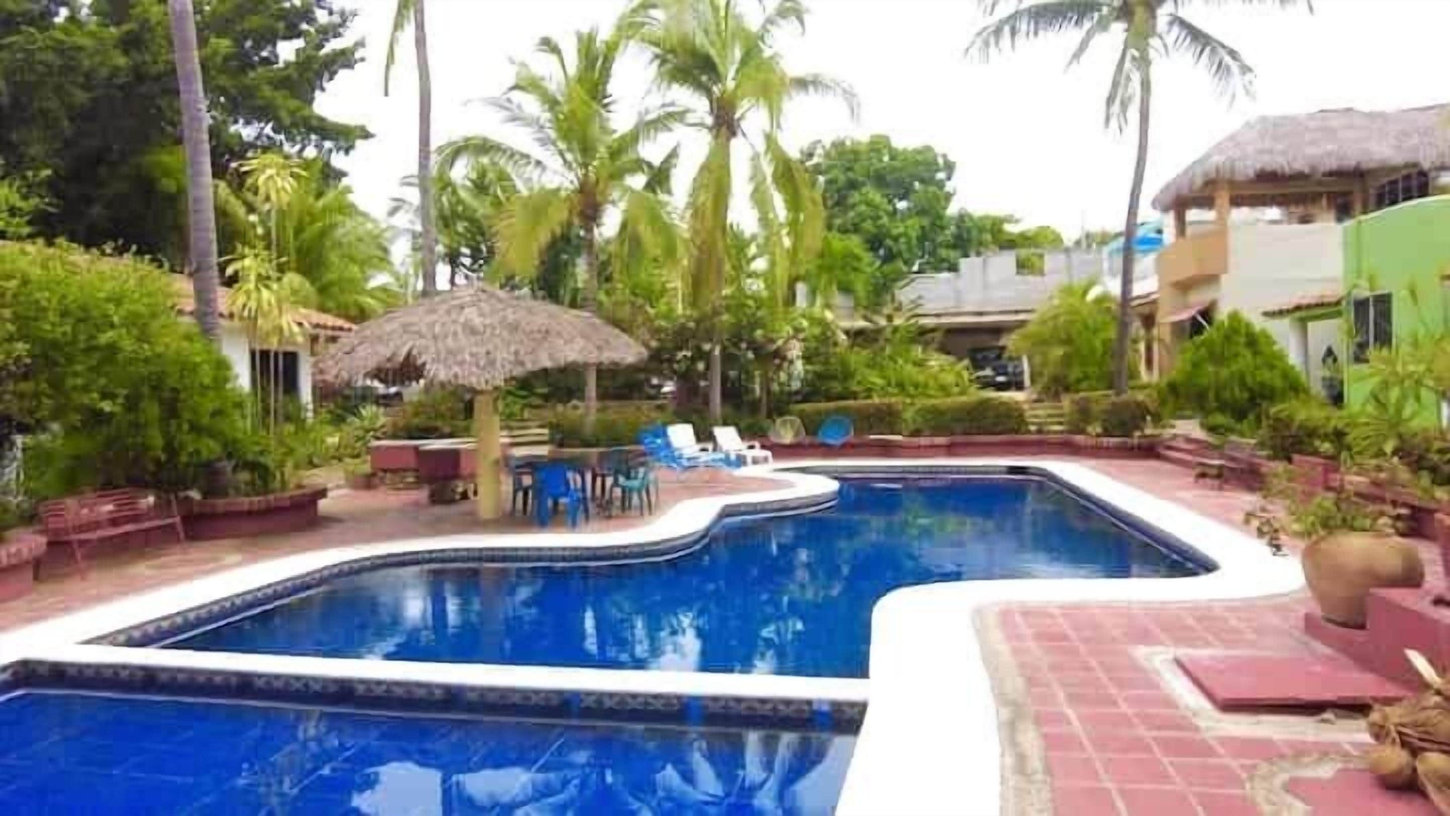 Una piscina con borde de azulejos, rodeada de palmeras y una estructura de techo de paja.