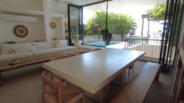 A modern living room with a white sofa, a dining table, and a view of a swimming pool through large glass doors. - Minimalist villa w/ private pool (Puerto Escondido)