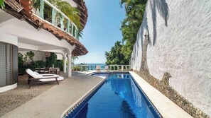 Une piscine avec des chaises longues, une vue sur la mer et un bâtiment blanc doté d’un toit décoratif.