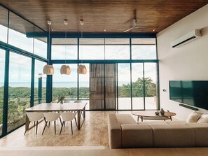 A spacious room with a large window, a dining area with a table and chairs, a sofa, and a flat-screen TV mounted on the wall. - Boutique Villa with Pool and Ocean View in Santa Teresa, Costa Rica (Santa Teresa)