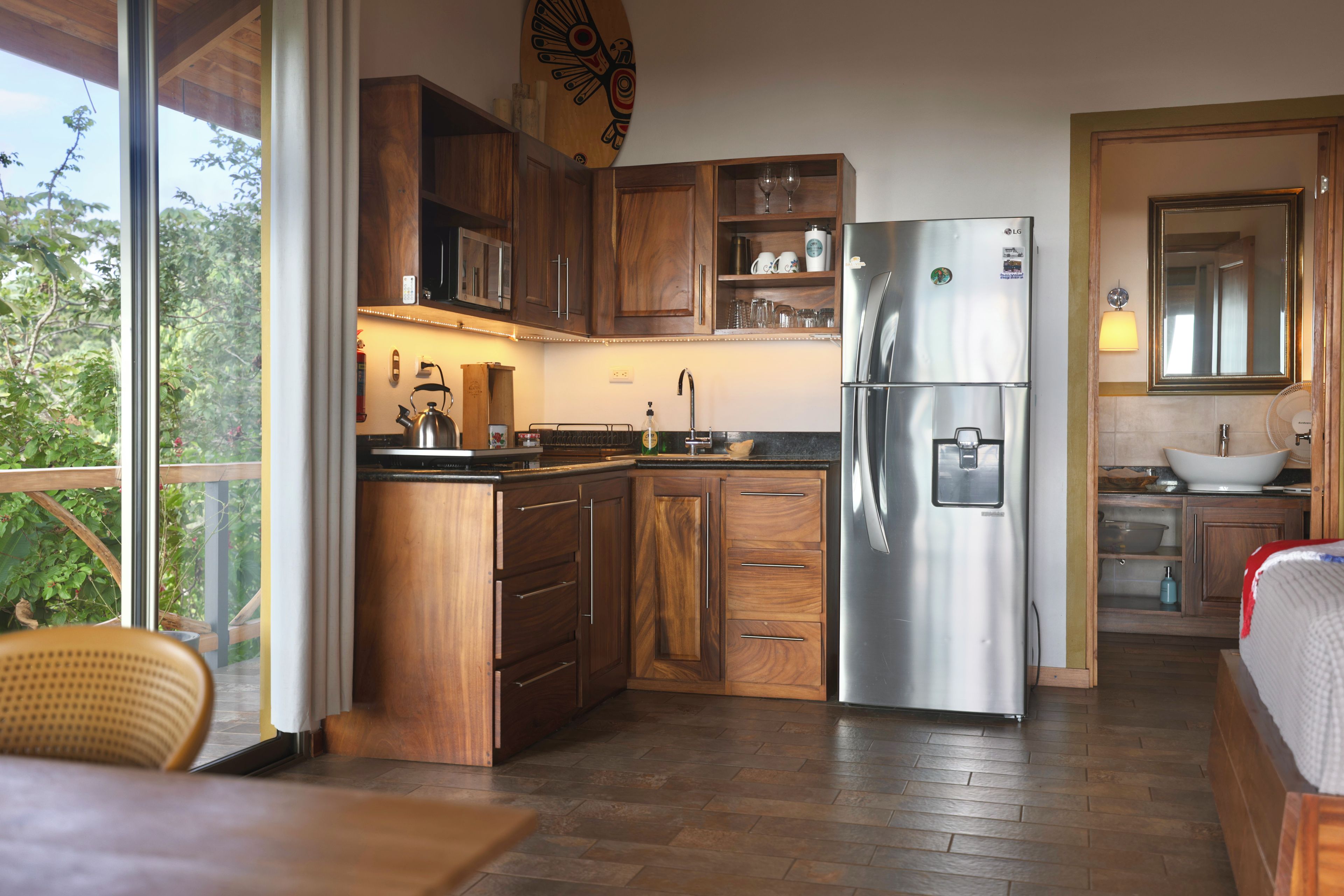 Cocina compacta con armarios de madera, refrigerador de acero inoxidable y ventana con vista a la vegetación.