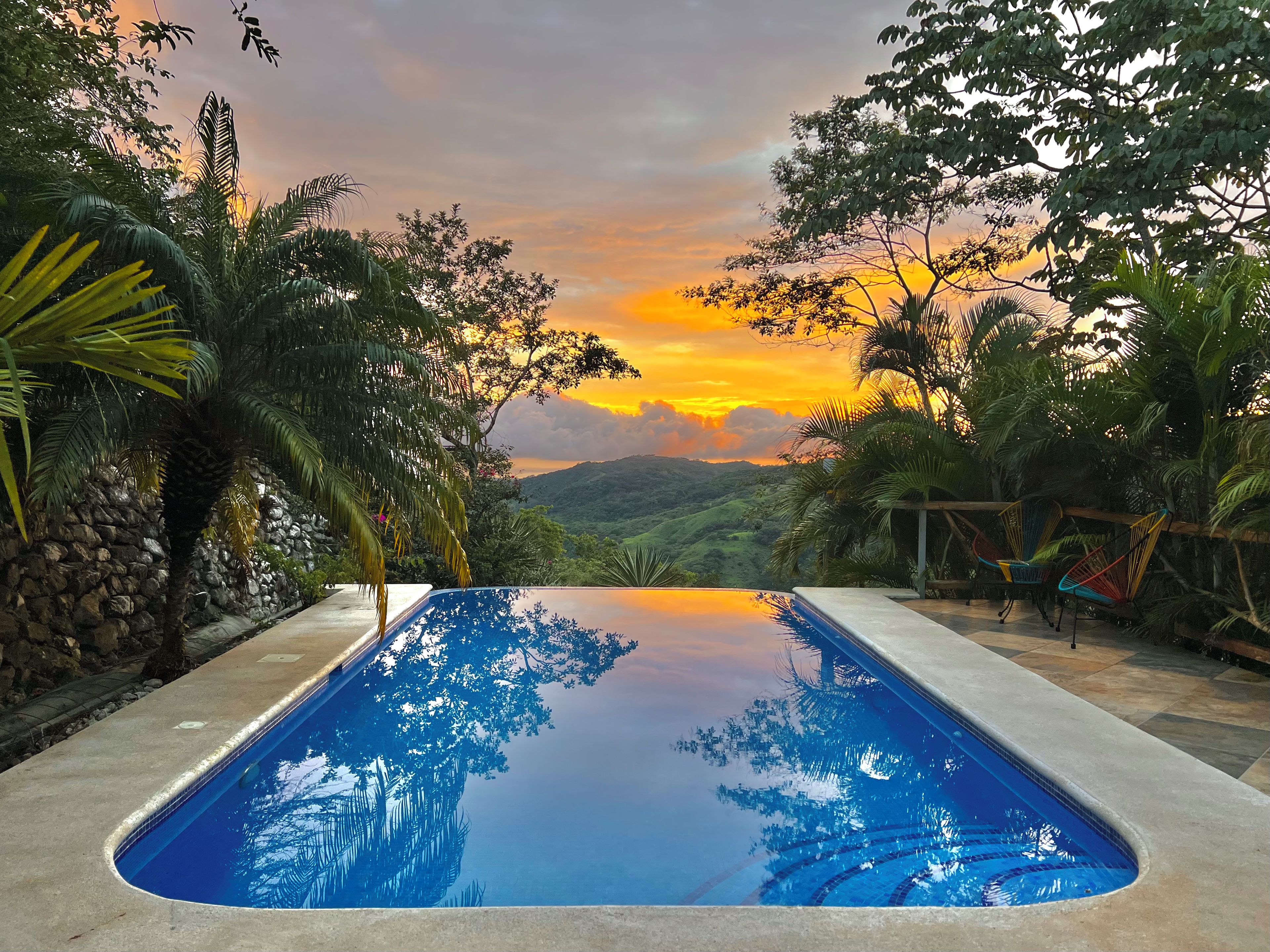 A swimming pool with a view of a sunset over a hilly landscape, surrounded by tropical vegetation and stone walls.
