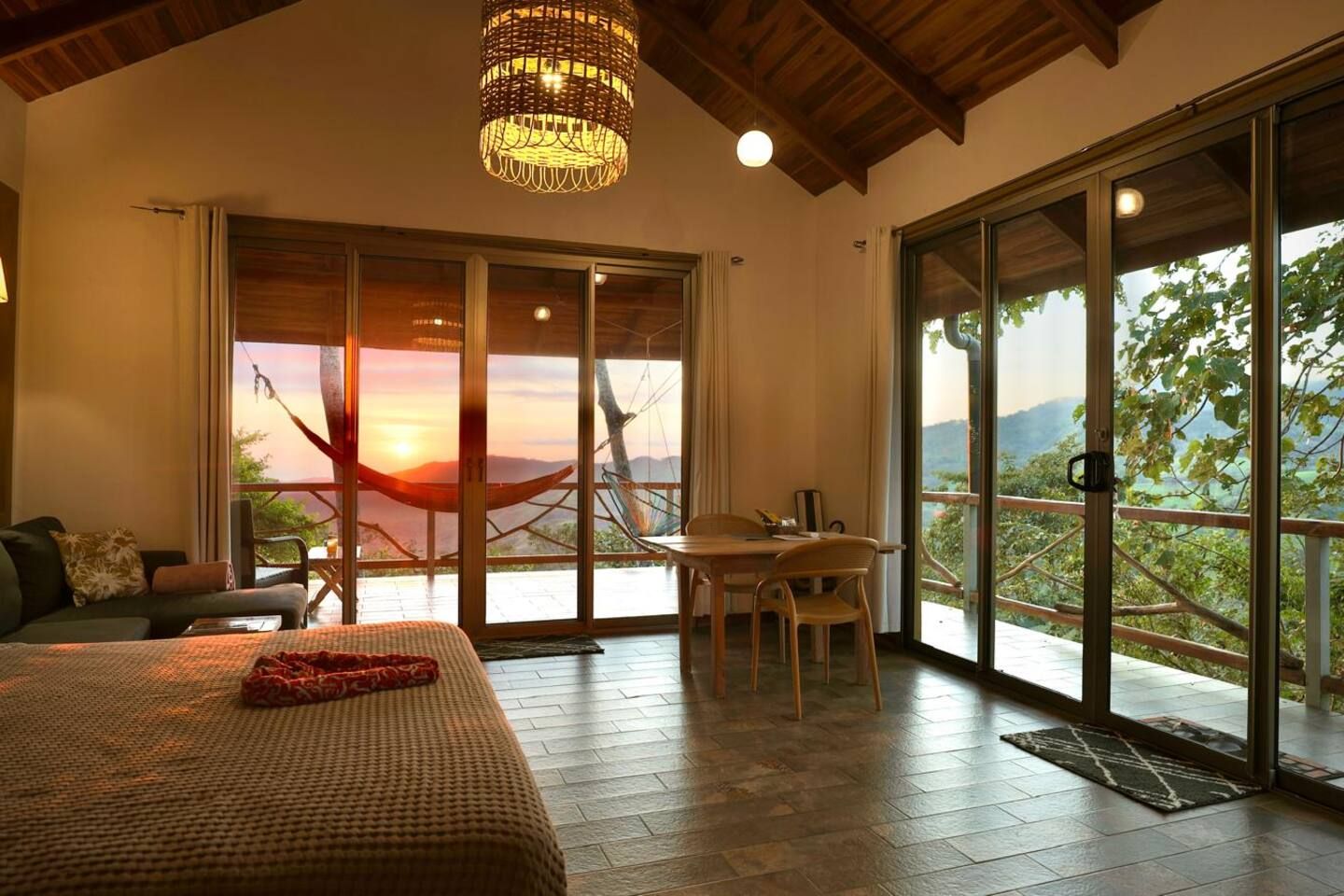 A bedroom with a large bed, a dining area with a table and chairs, and a view of the outdoors through large sliding doors.