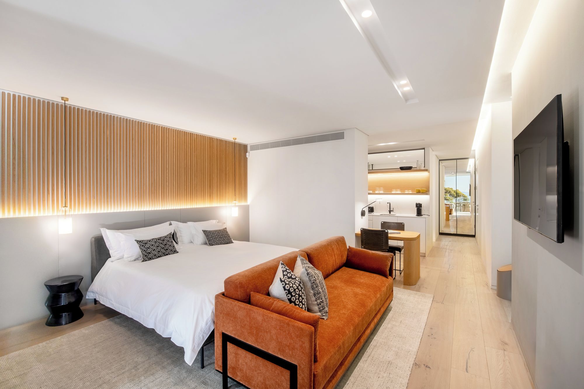 A modern hotel room with a bed, sofa, and a view of the kitchen and balcony.
