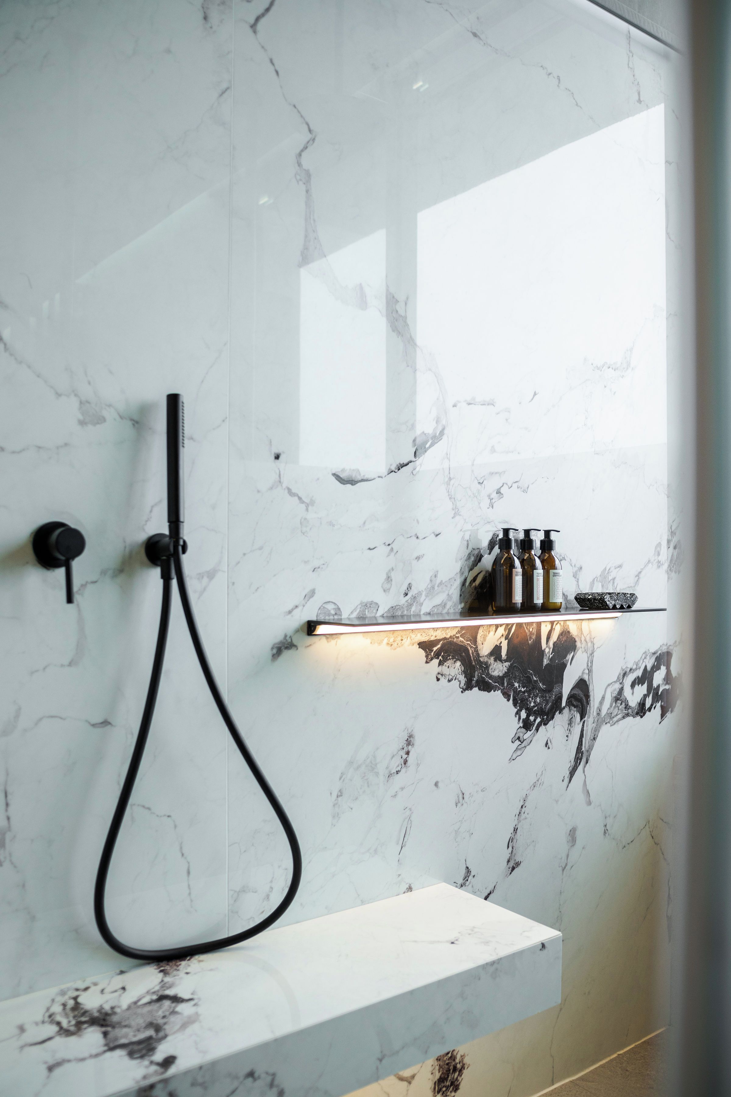 A modern bathroom with a marble wall, a handheld showerhead, and a shelf with toiletries.