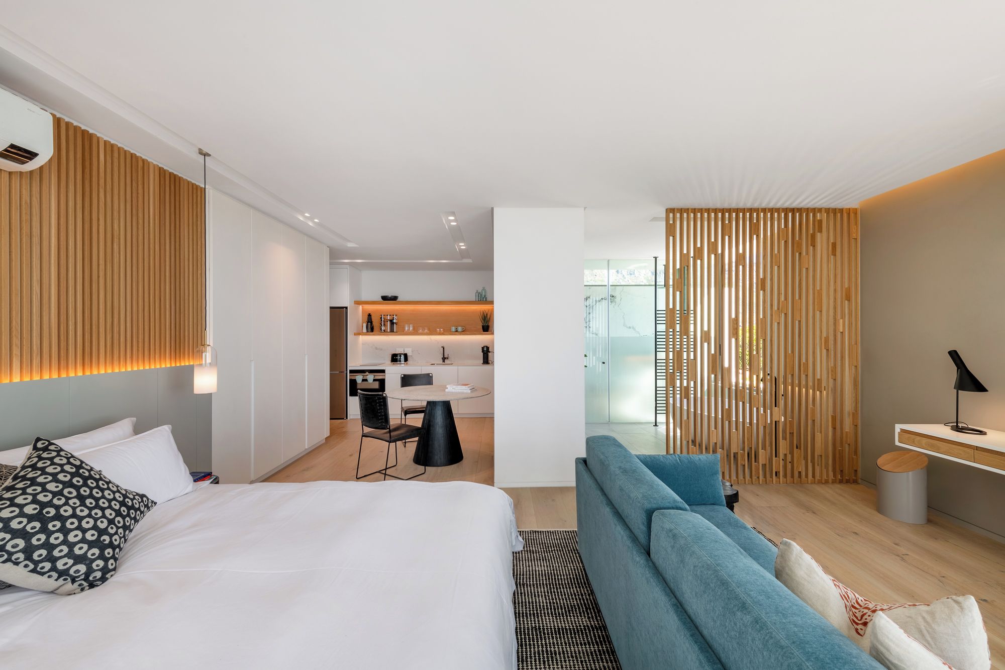 A modern hotel room with a bed, a seating area, a kitchenette, and a view through a glass door.