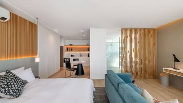 A modern hotel room with a bed, a seating area, a kitchenette, and a view through a glass door.