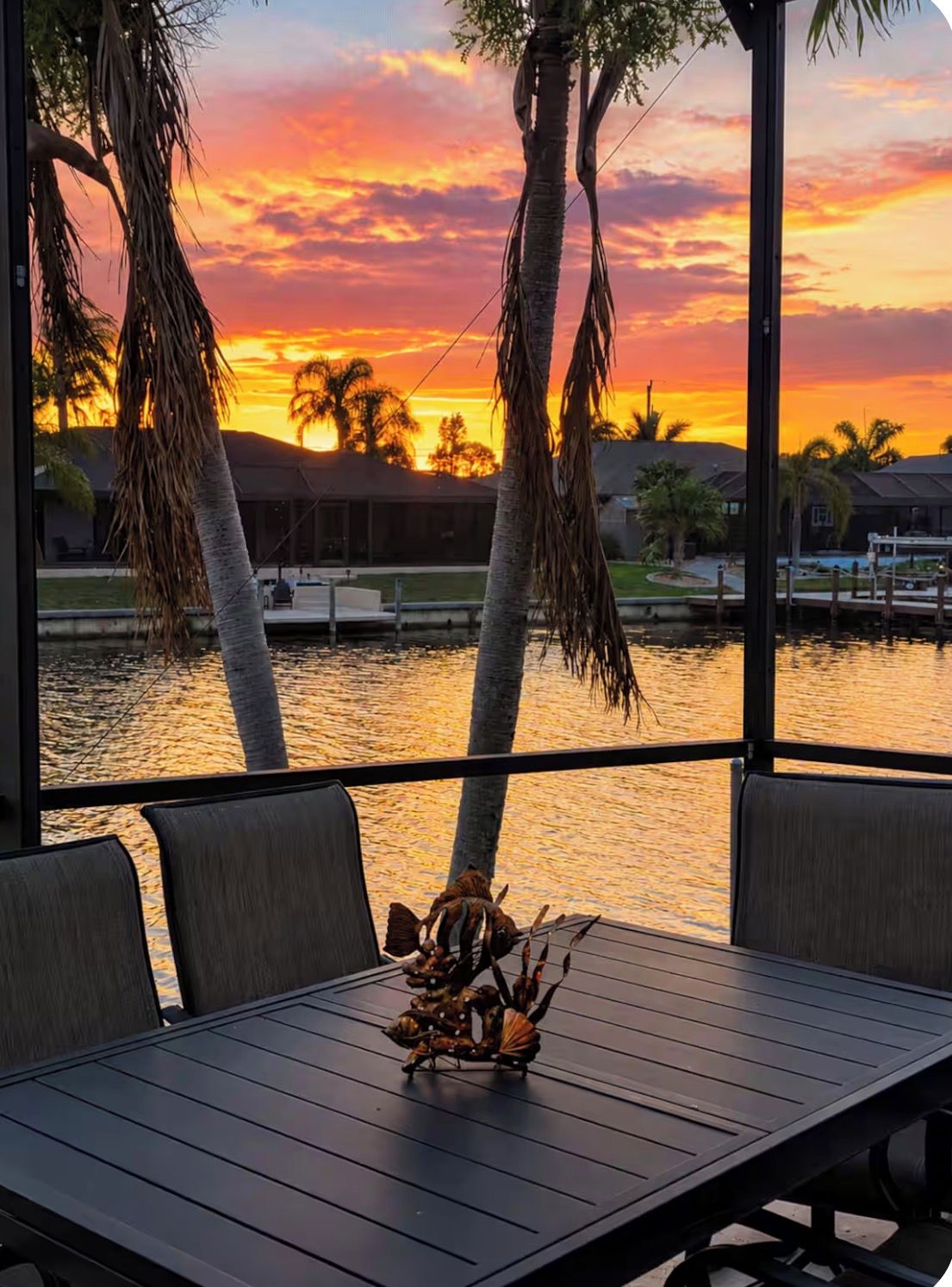 A table with chairs and a decorative centerpiece, overlooking a body of water with palm trees and a sunset.