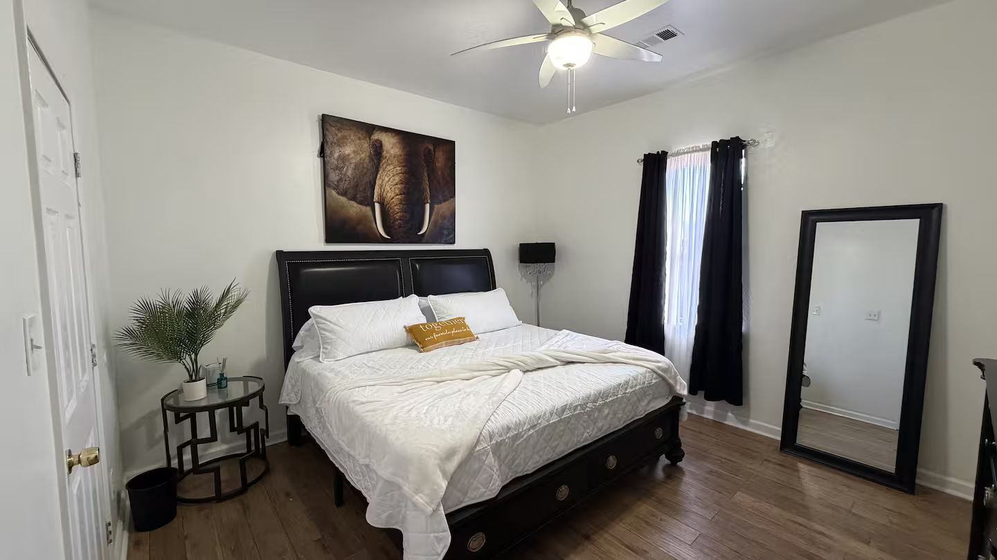 Un dormitorio con cama, ventilador de techo, ventana con cortinas, una puerta y una obra de arte colgada en la pared.