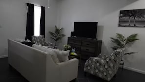 Ein Wohnzimmer mit Sofa, Sessel, Fernseher und Zimmerpflanzen.