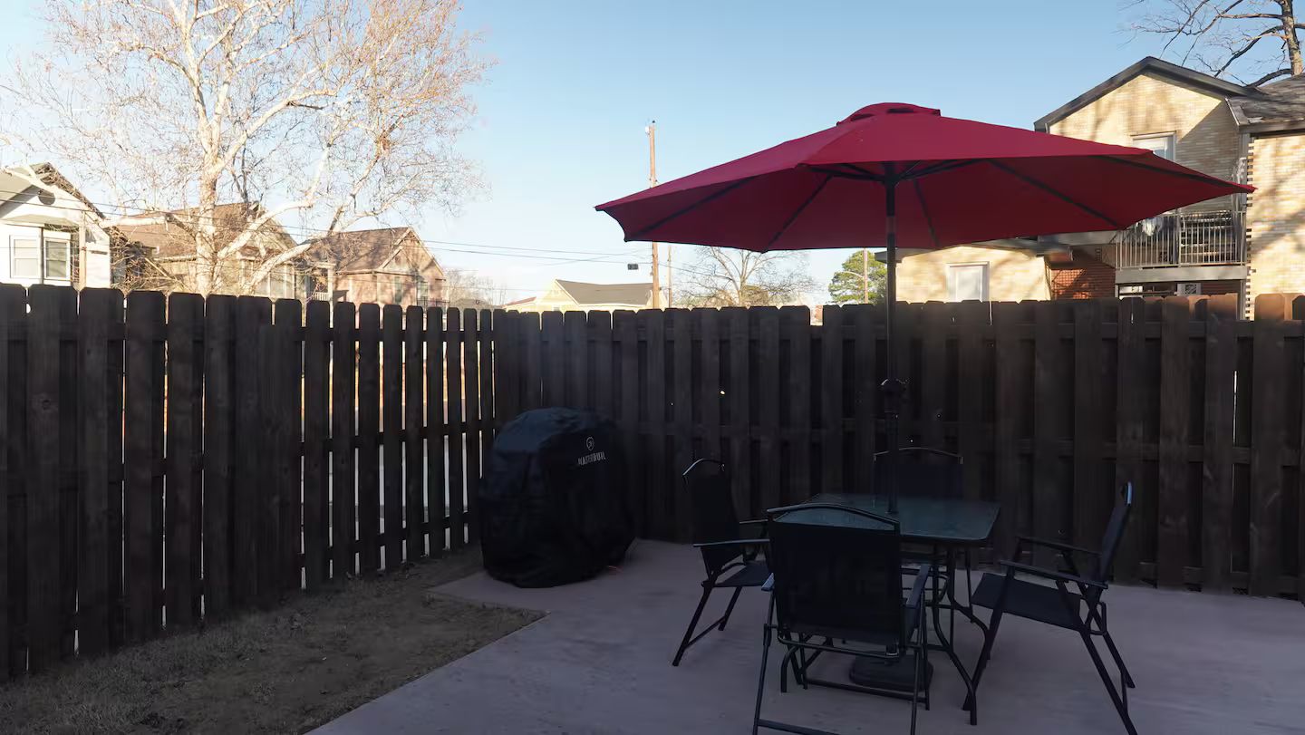 Eine Terrassenanlage im Freien mit einem roten Sonnenschirm, einem überdachten Grill und einem Tisch mit Stühlen.