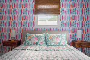 Quarto com cama, almofadas florais, janela com persianas e papel de parede estampado.