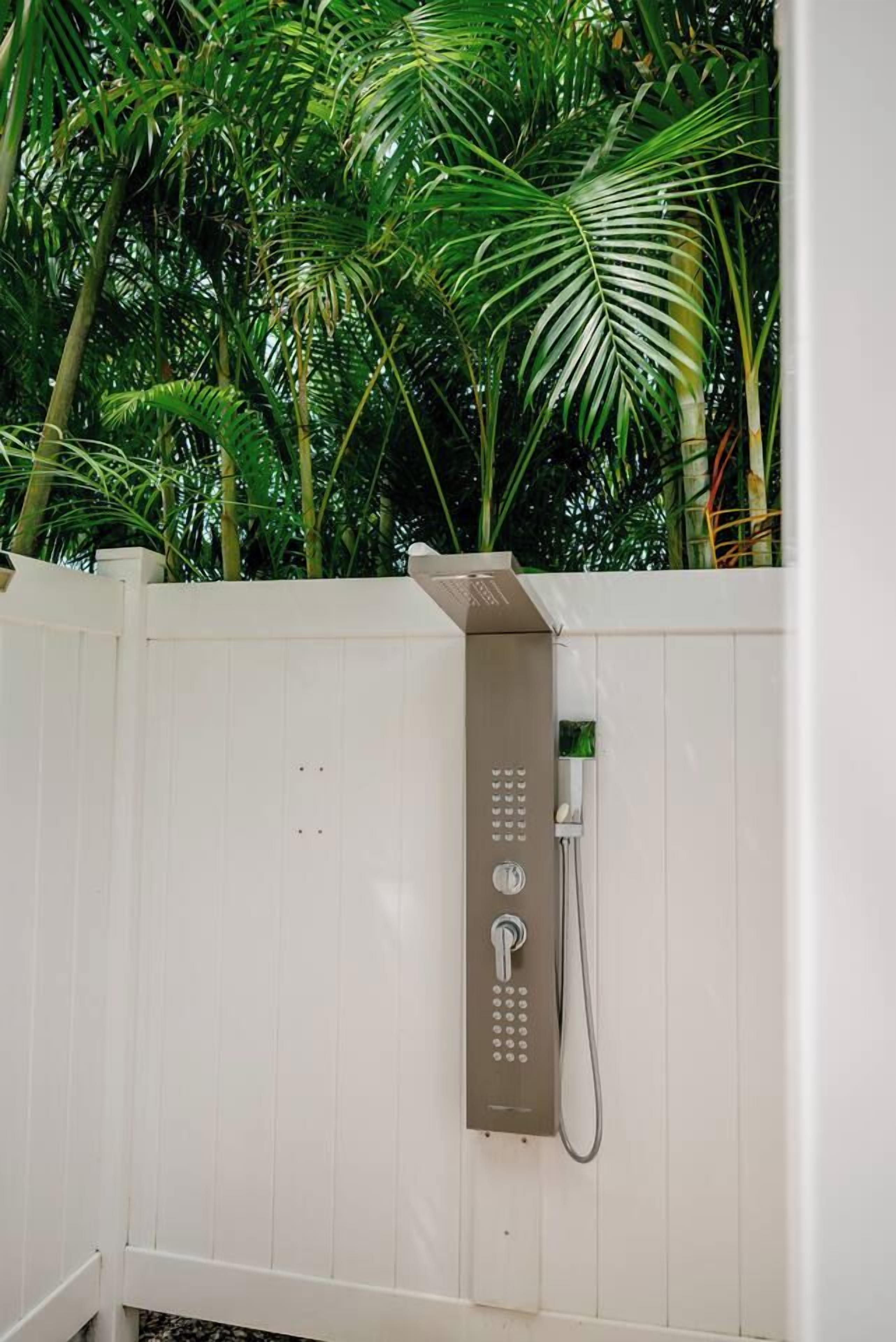 Une cabine de douche extérieure blanche, dotée d’un panneau de commande, et entourée d’un luxuriant feuillage verdoyant.