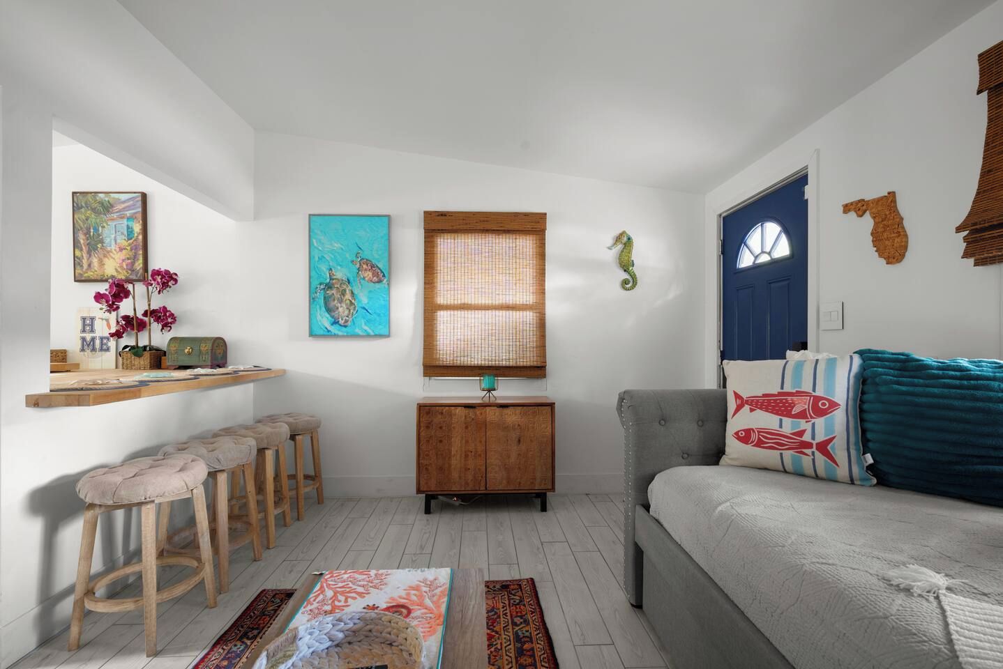 A compact living space with a sofa, a wooden cabinet, a small dining area with stools, and a window with a bamboo blind.