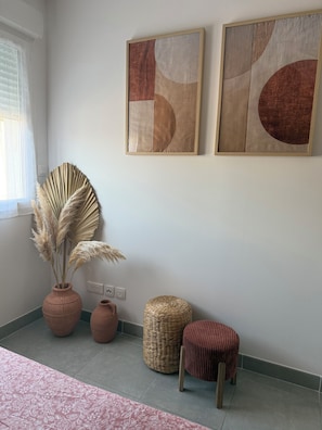 A room with a bed, a window with curtains, a wall-mounted artwork, a vase with dried plants, and a woven basket. - Cozy ground-floor apartment, private parking (Fos-sur-Mer)