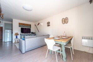 Ein modernes Wohnzimmer mit grauem Sofa, einem Holzesstisch mit weißen Stühlen, einem Fernseher und einer Küche im Hintergrund.
