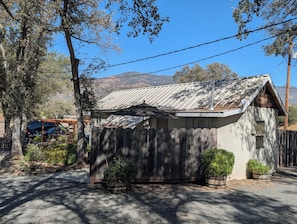 A small wooden cabin with a metal roof, surrounded by trees and a gravel driveway.