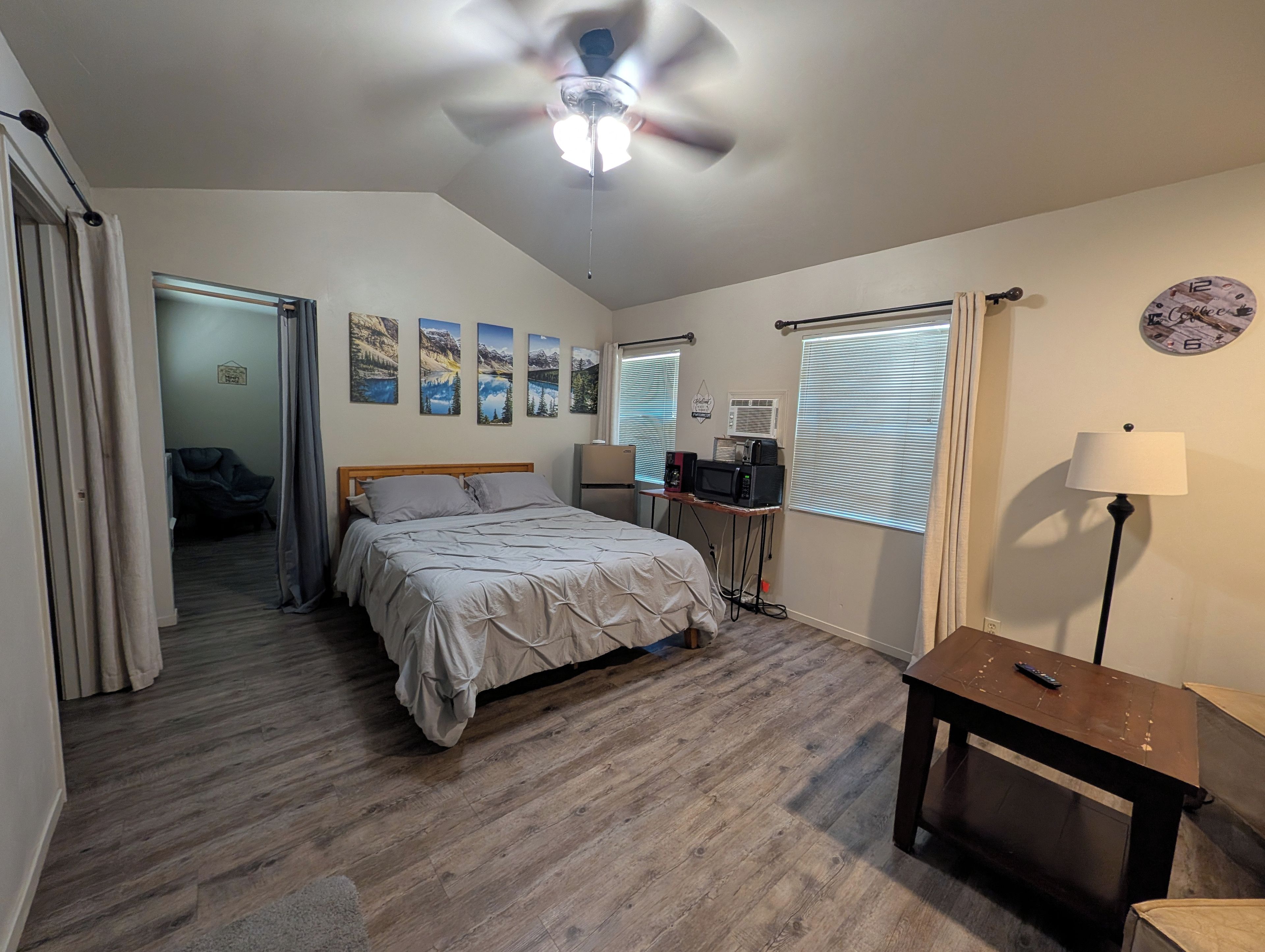A bedroom with a bed, a ceiling fan, a desk with a lamp, a clock, and a window with blinds.