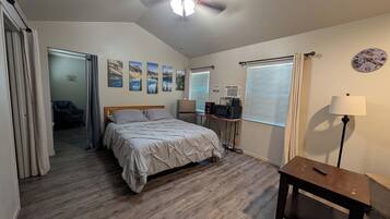 A bedroom with a bed, a ceiling fan, a desk with a lamp, a clock, and a window with blinds.