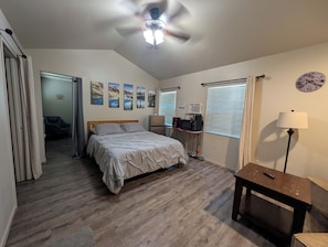A bedroom with a bed, a ceiling fan, a desk with a lamp, a clock, and a window with blinds.